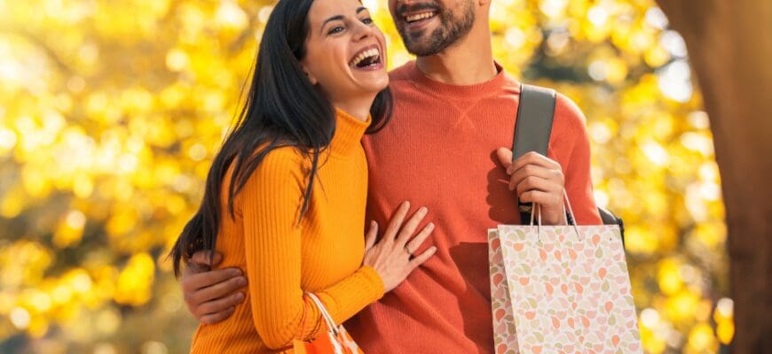 A man and a woman wlaking outside holding shopping bags with fall leaves in the background